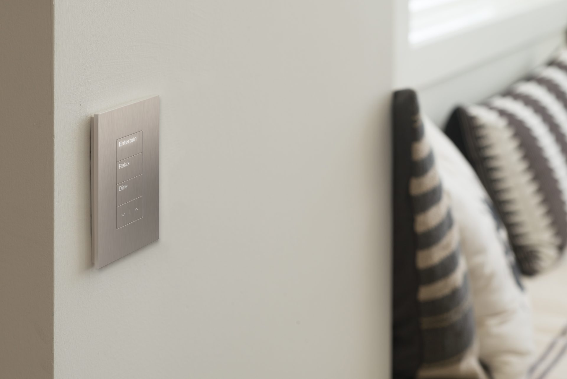 Close-up of a single engraved keypad on a wall, one button lit, with a calm evening room softly out of focus behind it.